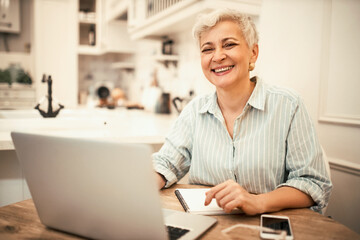 Cheerful middle aged woman in casual shirt laughing, being in positive mood, using portable...