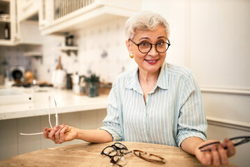 Indoor image of emotional retired woman with short gray hair sitting at wooden table with many eyeglasses, trying different frames enjoying online shopping, choosing best pair of spectacles