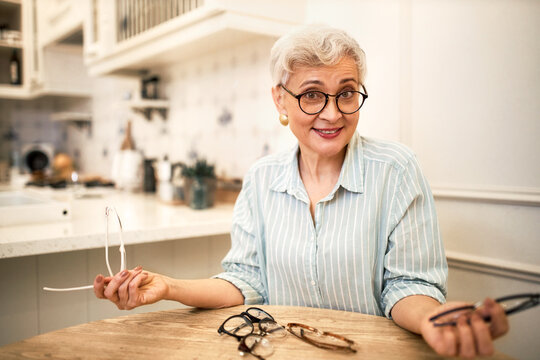 Indoor image of emotional retired woman with short gray hair sitting at wooden table with many eyeglasses, trying different frames enjoying online shopping, choosing best pair of spectacles