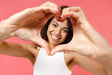 Young happy smiling romantic girlfriend lovely african american woman 20s wearing casual white tank shirt showing shape heart with hands looking through heart-shape sign isolated on pink background.