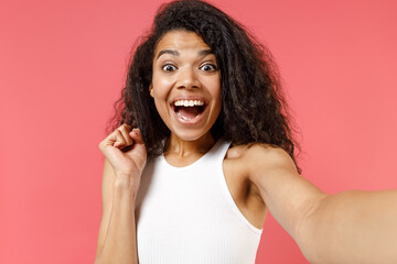 Close up young overjoyed african american woman 20s wearing white tank shirt doing selfie shot on mobile phone talk by video do winner gesture clench fist isolated on pink background studio portrait