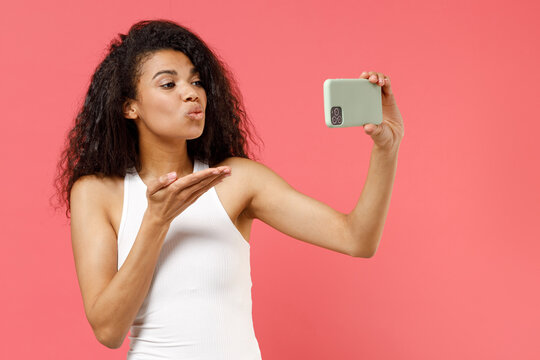 Young romantic happy girlfriend lovely african american woman in white tank shirt do selfie shot on mobile phone blowing air kiss isolated on pink background studio portrait. People lifestyle concept.