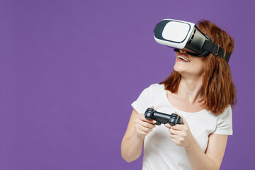 Young excited fun redhead caucasian woman 20s wearing white basic casual t-shirt playing pc game with joystick console watching in vr headset pc gadget isolated on dark violet color background studio