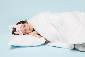 Young sleeping calm woman in pajamas jam sleep eye mask rest relaxing at home lying lies wrap covered under blanket duvet on pillow isolated on pastel blue background. Good mood night bedtime concept.