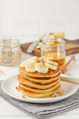 A stack of oatmeal banana pancakes with slices of fresh bananas, walnuts and honey on top with cup of tea on a white wooden background. A healthy breakfast. Copy space.
