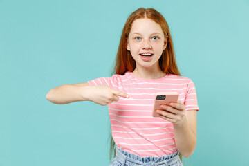 Little fun redhead kid girl 12-13 years old in pink striped t-shirt point index finger on mobile cell phone chatting isolated on pastel blue background studio. Children lifestyle childhood concept