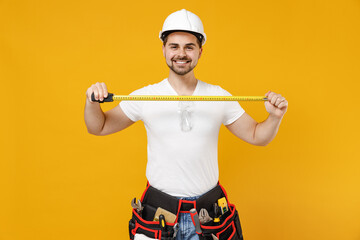 Young employee handyman man in protective helmet hardhat hold measurement meter tape isolated on yellow background studio portrait Instrument accessories renovation apartment room Repair home concept