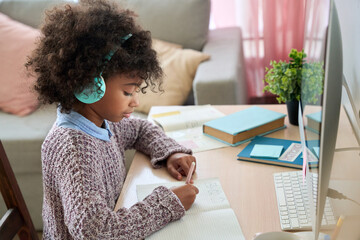 Concentrated preteen black African American girl wear headphones sitting at table writing listening to tutor online virtual lesson studying at home doing task. Remote distant homeschooling concept.
