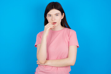 Nervous puzzled young beautiful Caucasian woman wearing pink T-shirt over blue wall opens mouth from surprise, reacts on sudden news.