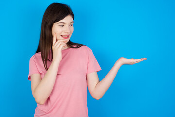 Funny young beautiful Caucasian woman wearing pink T-shirt over blue wall holding open palm new product. I wanna buy it!