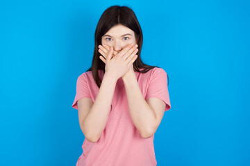 young beautiful Caucasian woman wearing pink T-shirt over blue wall shocked covering mouth with hands for mistake. Secret concept.