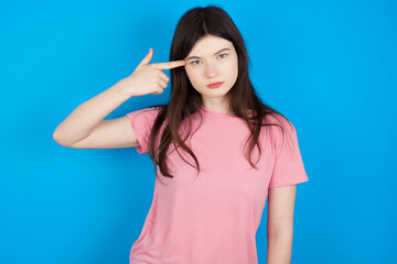 Unhappy young beautiful Caucasian woman wearing pink T-shirt over blue wal makes suicide gesture and imitates gun with hand, curves lips keeps two fingers on temple, shoots, being tired of everything,