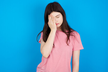 Tired overworked young beautiful Caucasian woman wearing pink T-shirt over blue wall has sleepy expression, gloomy look, covers face with hand, has eyes shut, gasps from tiredness, fatigue after party