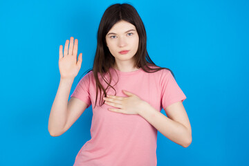 young beautiful Caucasian woman wearing pink T-shirt over blue wall Swearing with hand on chest and open palm, making a loyalty promise oath