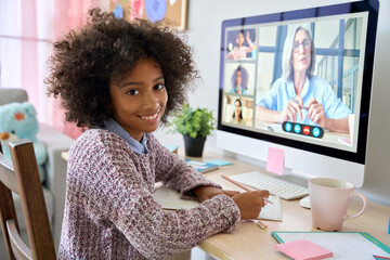 Smiling African American teen pupil looking at camera writing homework at desk with video class with teacher tutor on desktop computer at home. Remote distant homeschooling education concept.