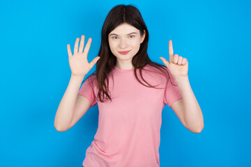 young beautiful Caucasian woman wearing pink T-shirt over blue wall showing and pointing up with fingers number seven while smiling confident and happy.