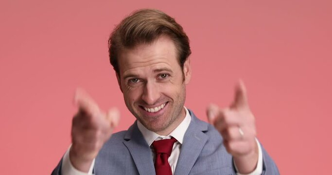 attractive elegant businessman in suit pointing fingers and smiling while standing and posing on pink background in studio