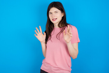 young beautiful Caucasian woman wearing pink T-shirt over blue wall afraid and terrified with fear expression stop gesture with hands, shouting in shock. Panic concept.