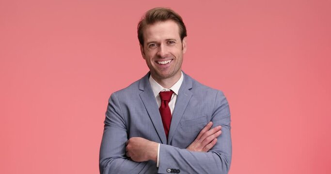 elegant smiling man in blue suit looking down, being happy and crossing arms while nodding on pink background in studio