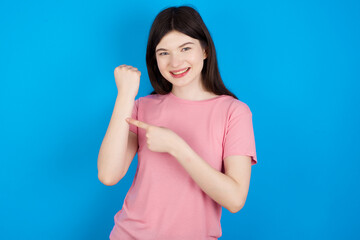 young beautiful Caucasian woman wearing pink T-shirt over blue wall In hurry pointing to wrist watch, impatience, looking at the camera with relaxed expression