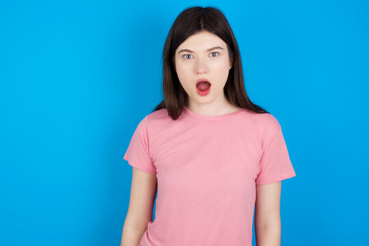 Oh my God. Surprised young beautiful Caucasian woman wearing pink T-shirt over blue wall stares at camera with shocked expression exclaims with unexpectedness,