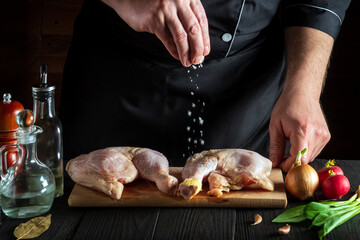 Chef or cook adds salt to the chicken legs. Preparation for grilling with chicken leg in the kitchen of a restaurant or cafe on a table with vegetables and spices