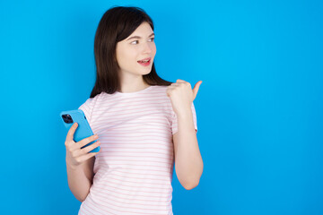 young beautiful Caucasian woman wearing stripped T-shirt over blue wall using and texting with smartphone  pointing and showing with thumb up to the side with happy face smiling