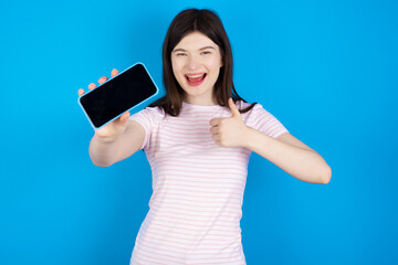 Portrait young beautiful Caucasian woman wearing stripped T-shirt over blue wall holding in hands cell showing giving black screen thumb up