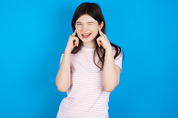 Happy young beautiful Caucasian woman wearing stripped T-shirt over blue wall ignores loud music and plugs ears with fingers asks to turn off sound