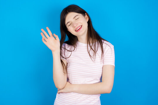 Overjoyed successful young beautiful Caucasian woman wearing stripped T-shirt over blue wall raises palm and closes eyes in joy being entertained by friends