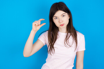 young beautiful Caucasian woman wearing stripped T-shirt over blue wall purses lip and gestures with hand, shows something very little.