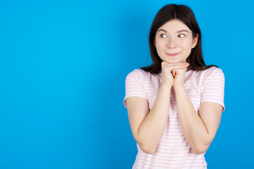 Curious young beautiful Caucasian woman wearing stripped T-shirt over blue wall keeps hands under chin bites lips and looks with interest aside.