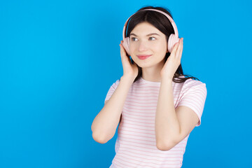 young beautiful Caucasian woman wearing stripped T-shirt over blue wall wears stereo headphones listens music concentrated aside. People hobby lifestyle concept