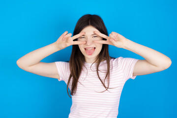 Obraz na płótnie Canvas Cheerful positive young beautiful Caucasian woman wearing stripped T-shirt over blue wall shows v-sign near eyes open mouth