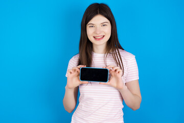 Cheerful cheery content young beautiful Caucasian woman wearing stripped T-shirt over blue wall holding in hands device hobby smm post blog
