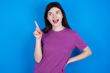 young beautiful Caucasian woman wearing purple T-shirt over blue wall holding finger up having idea and posing