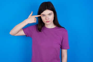 Unhappy young beautiful Caucasian woman wearing purple T-shirt over blue  makes suicide gesture and imitates gun with hand, curves lips keeps two fingers on temple, shoots, being tired of everything,