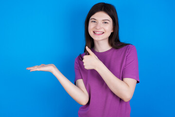 young beautiful Caucasian woman wearing purple T-shirt over blue wall Showing palm hand and doing ok gesture with thumbs up, smiling happy and cheerful.