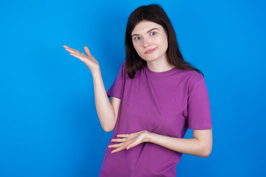 young beautiful Caucasian woman wearing purple T-shirt over blue wall pointing aside with both hands showing something strange and saying: I don't know what is this. Advertisement concept.