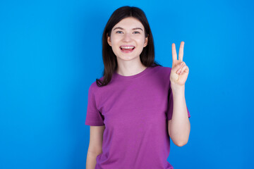 young beautiful Caucasian woman wearing purple T-shirt over blue wall showing and pointing up with fingers number two while smiling confident and happy.