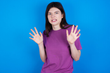 young beautiful Caucasian woman wearing purple T-shirt over blue wall afraid and terrified with fear expression stop gesture with hands, shouting in shock. Panic concept.