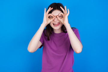 young beautiful Caucasian woman wearing purple T-shirt over blue wall doing ok gesture like binoculars sticking tongue out, eyes looking through fingers. Crazy expression.