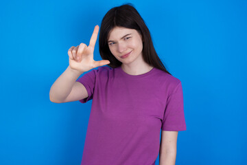 Fototapeta premium young beautiful Caucasian woman wearing purple T-shirt over blue wall making fun of people with fingers on forehead doing loser gesture mocking and insulting.
