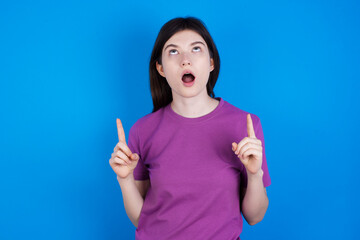 young beautiful Caucasian woman wearing purple T-shirt over blue wall amazed and surprised looking up and pointing with fingers and raised arms.