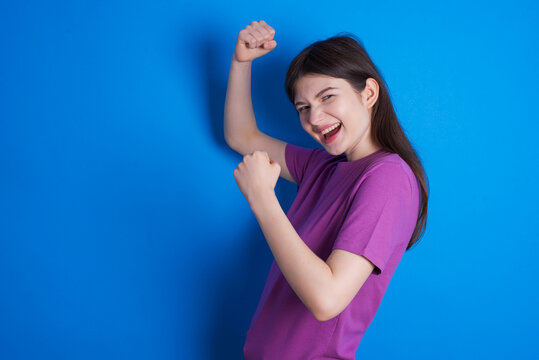 Profile photo of excited young beautiful Caucasian woman wearing purple T-shirt over blue wall raising fists celebrating black Friday shopping