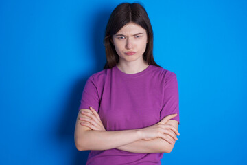 Obraz premium Gloomy dissatisfied young beautiful Caucasian woman wearing purple T-shirt over blue wall looks with miserable expression at camera from under forehead, makes unhappy grimace