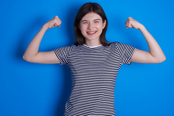 Strong powerful young beautiful Caucasian woman wearing pink jacket over blue wall toothy smile, raises arms and shows biceps. Look at my muscles!