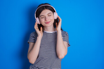 Pleased young beautiful Caucasian woman wearing stripped T-shirt over blue wall enjoys listening pleasant melody keeps hands on stereo headphones closes eyes. Spending free time with music