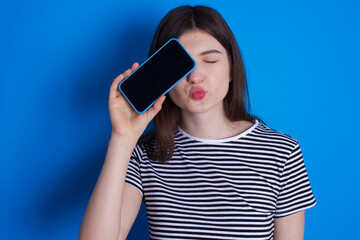 Adorable young beautiful Caucasian woman wearing stripped T-shirt over blue wall holding modern device covering eye with lips pouted