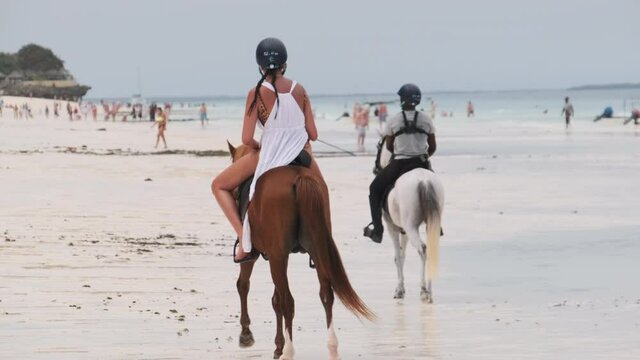 Horseback riding on a tropical beach along the coast of the Ocean. Woman horse riders move against the backdrop of exotic shore with tourists. Exotic sandy beach on a tropical coast, Africa, Tanzania
